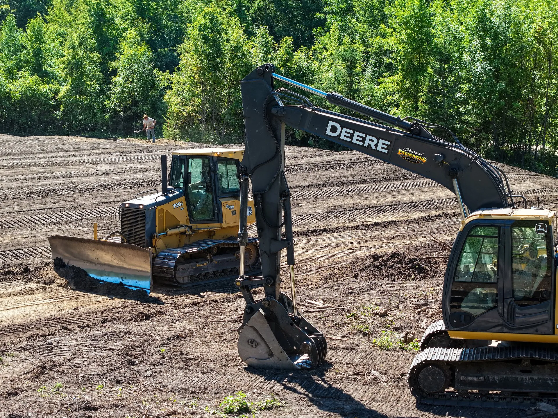 Low aerial view of a Shepard Excavating excavator grading a lakefront lot near Walker, Minnesota, with birch and pine trees behind it.